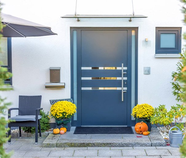 Modern house entrance with grey door, potted yellow flowers, pumpkins, and outdoor seating.