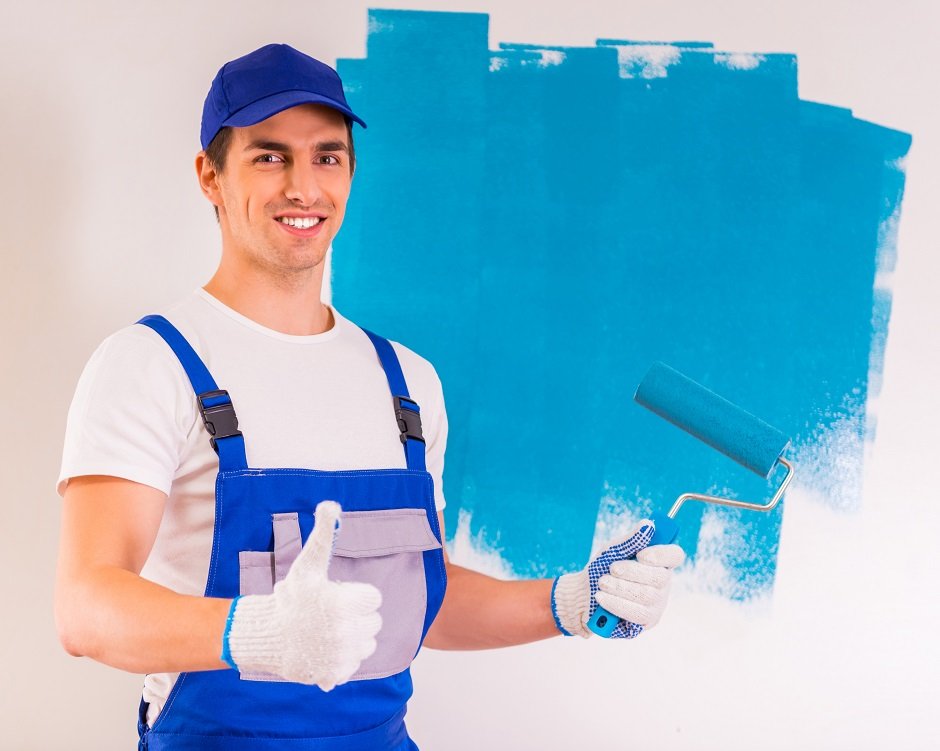Smiling painter in blue overalls giving a thumbs up while painting a wall light blue with a roller.